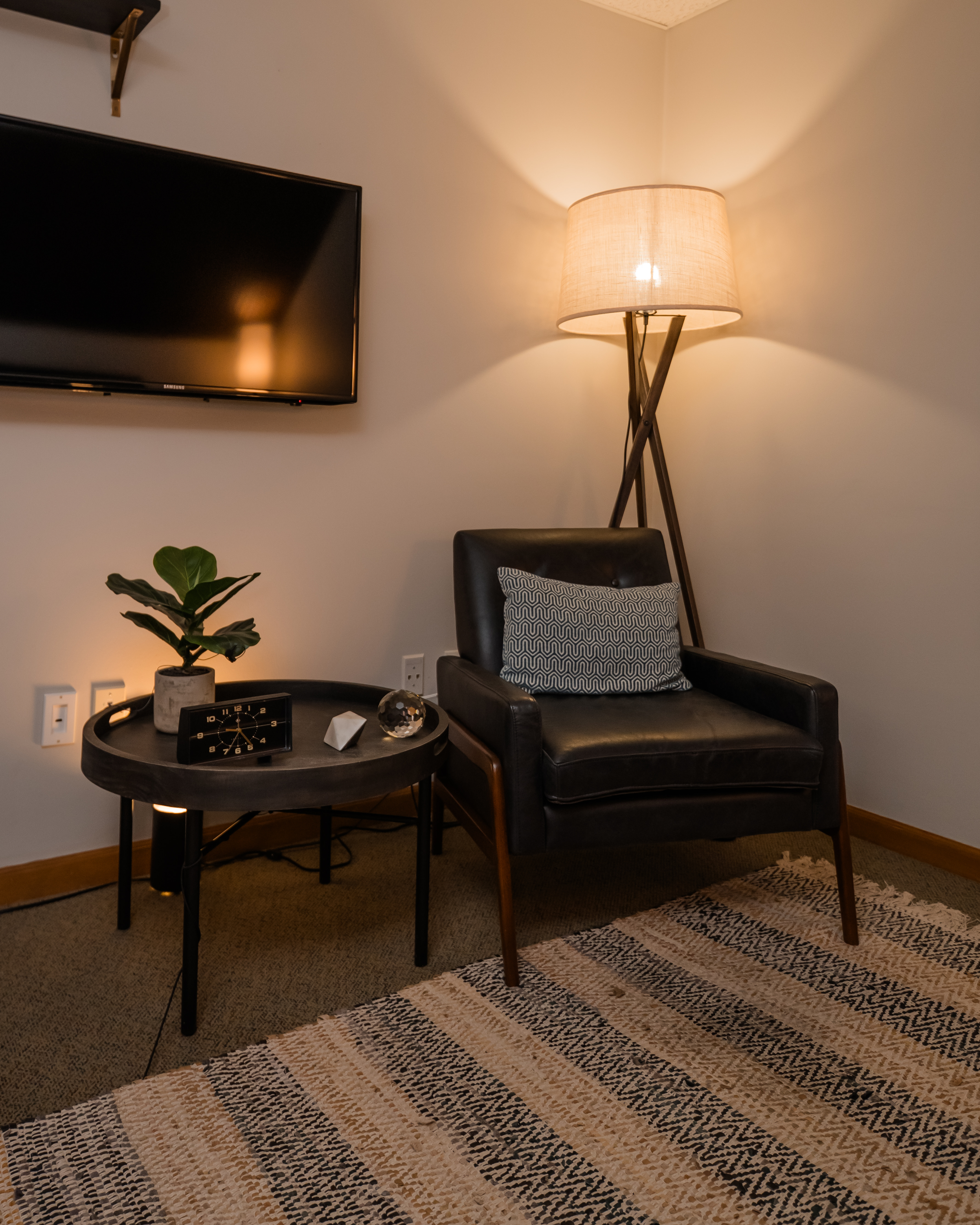 Dark leather chair with round side table set underneath a wall mounted TV with lamp in background.